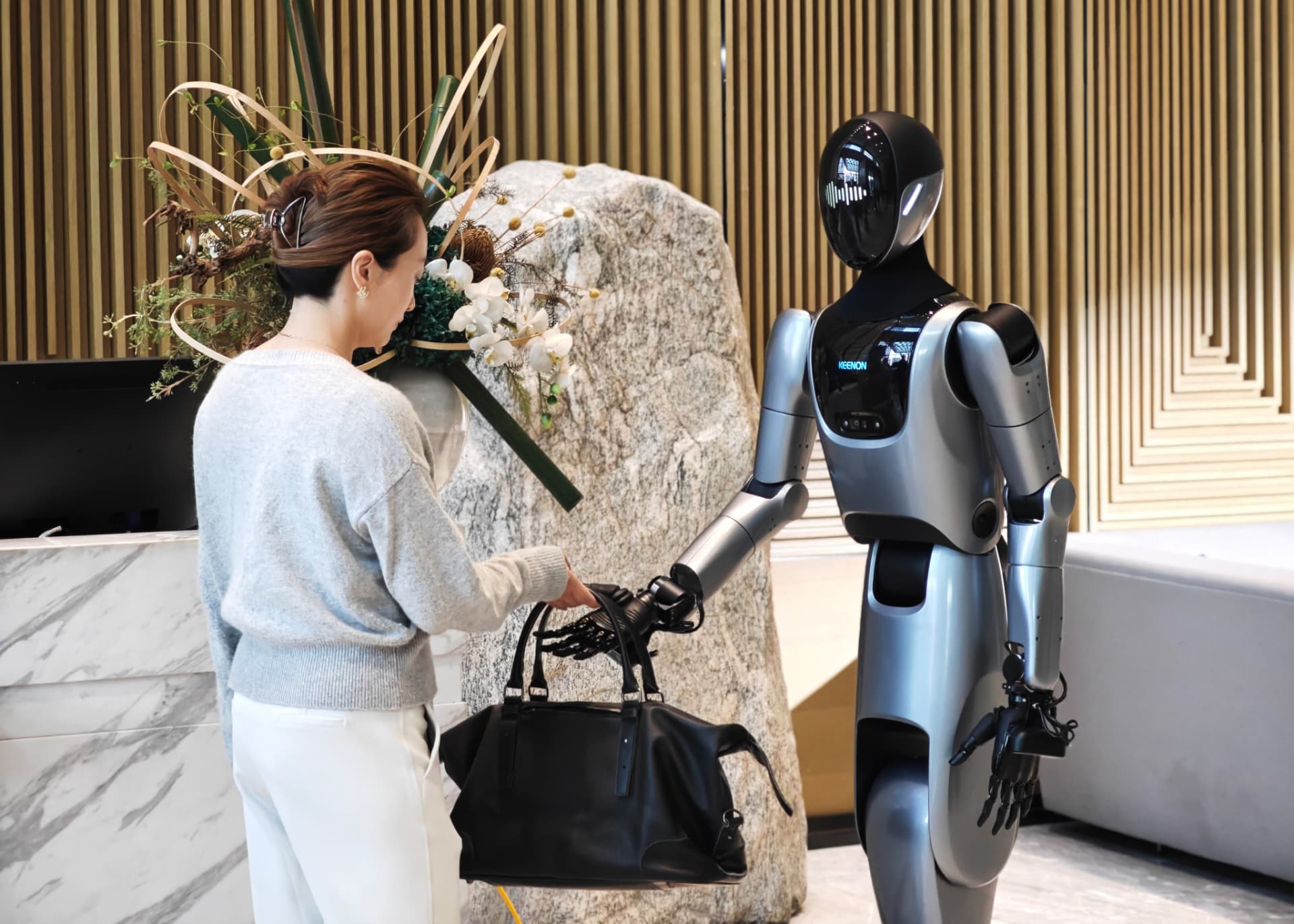 KEENON XMAN-R1 humanoid service robot handing a guest their bag beside a hotel reception desk